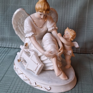 May include: A porcelain angel figurine with a seated woman and a small cherub. The woman wears a white dress with gold trim and holds a harp. The cherub has small wings and is reaching towards the woman. The figurine has a gold trim and is on an oval base.