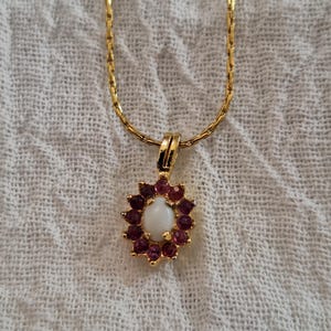 May include: A gold-tone chain necklace featuring a pendant with a central oval white cabochon stone surrounded by a halo of small, round, deep pink-red stones set in a scalloped gold-tone metal frame with a simple bail attachment.