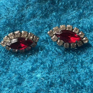 May include: Two vintage earrings with a marquise-shaped, deep red center stone surrounded by a halo of small, clear rhinestones. The earrings are set against a textured, turquoise background.