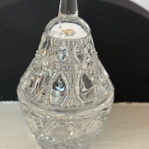 May include: A clear, glass jar with a lid. The jar has a textured, patterned design and a small, clear knob on top. The jar is likely used for storing small items or as a decorative piece.