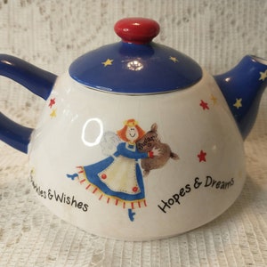 May include: A ceramic teapot with a white body, blue handle and spout, and a blue lid with a red knob. The teapot is decorated with a cartoon angel, stars, and the words "Sparkles & Wishes" and "Hopes & Dreams".