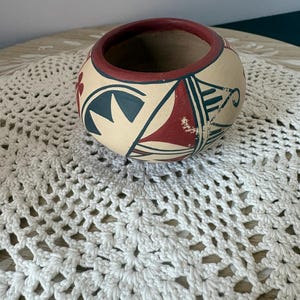 May include: A small, round, hand-painted ceramic bowl. The bowl features a cream-colored base with geometric designs in red, teal, and black. The rim is painted red. The bowl is approximately 4 inches in diameter.