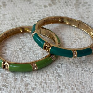 May include: Two gold-toned hinged bangles with green and emerald enamel and small clear crystals. The bracelets are on a white lace surface. The bangles are a fashion accessory.