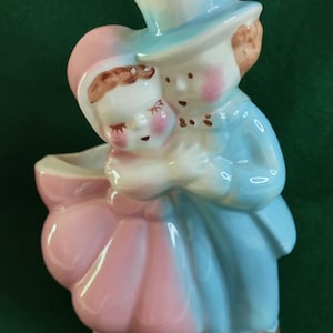May include: A ceramic planter shaped like a couple holding each other. The woman is wearing a pink dress and the man is wearing a blue suit and a top hat.