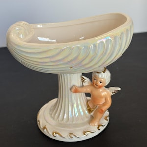 May include: A pearlescent white ceramic dish with a shell-like design, supported by a fluted column and a cherub figurine. The dish has a curved, boat-like shape. Gold accents adorn the base.