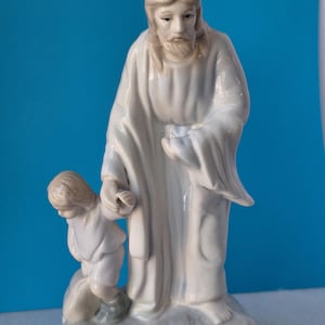 May include: A ceramic figurine depicting a figure in a white robe with long hair and a beard, holding the hand of a smaller figure. The figures are standing on a white base with a blue background.