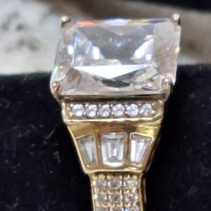 May include: A gold ring featuring a large rectangular center stone, a row of small round stones, three baguette-cut stones, and a band accented with multiple tiny round stones. The metal has a polished yellow finish and a multi-tiered design.