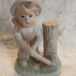 May include: A porcelain figurine of a young person in overalls and a hat, chopping wood with an axe. The figure is light pink and white, with a light green base and a small tree stump.