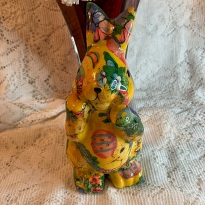 May include: A yellow ceramic Easter bunny figurine with a colorful floral and egg design. The bunny is standing on a white lace tablecloth.