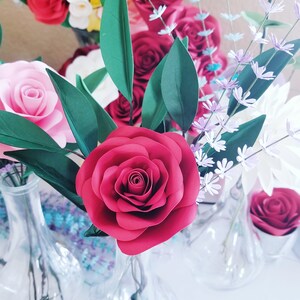 May include: A bouquet of paper flowers in a clear glass vase. The bouquet includes red, pink, and white roses, as well as small purple flowers.