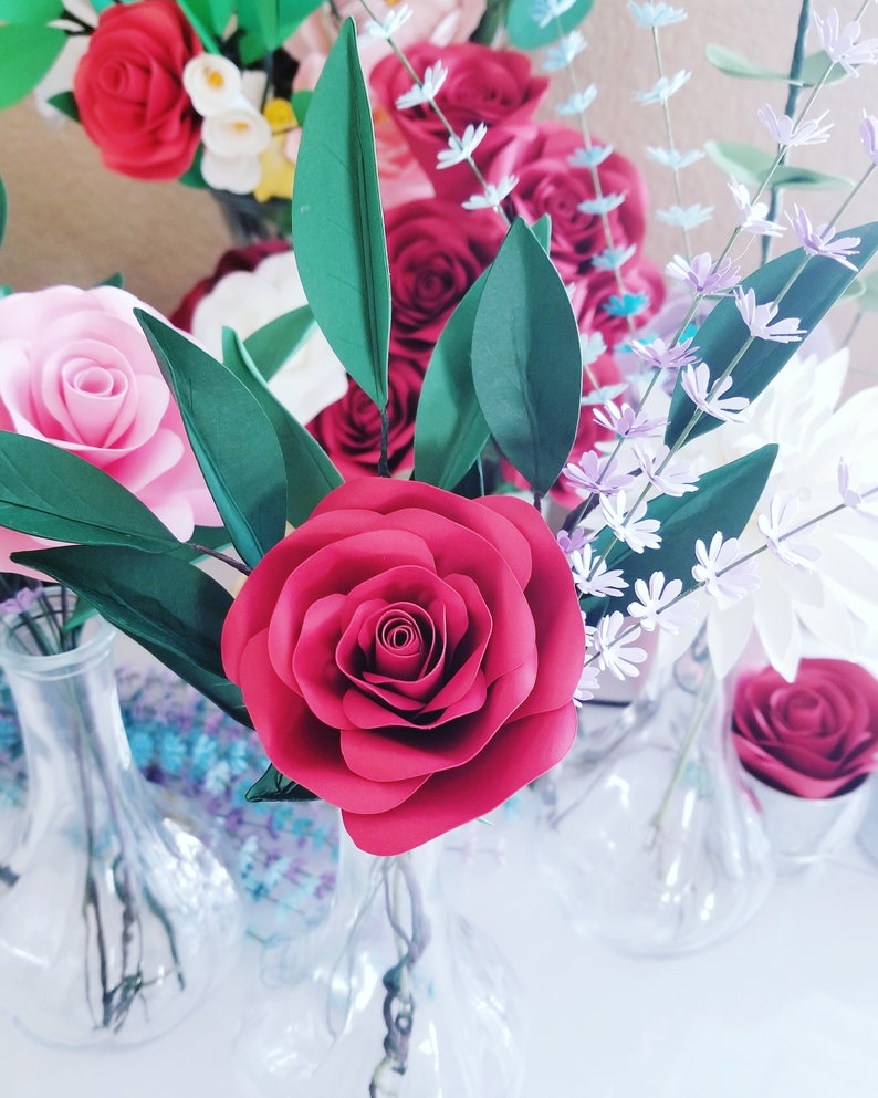 May include: A bouquet of paper flowers in a clear glass vase. The bouquet includes red, pink, and white roses, as well as small purple flowers.
