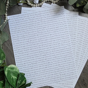 Könnte beinhalten: Ein Stapel weißer Papierbögen, bedruckt mit schwarzem Text aus der Bibel, speziell dem Buch Genesis, verziert mit einer Kette aus weißen Kunstperlen und grünen Blättern, die um die Ränder der Dokumente angeordnet sind.