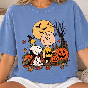 May include: A blue short-sleeve t-shirt featuring a graphic of Charlie Brown, Snoopy, and Woodstock in a Halloween scene. The design includes a large yellow moon, bats, a bare tree, orange pumpkins, and scattered autumn leaves on the ground.