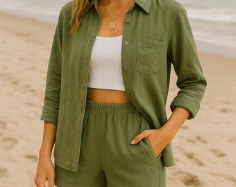 May include: A woman in an olive green linen outfit, comprising a shirt, shorts, and a white crop top, stands on a beach. She wears a gold pendant necklace. The background features the sea and sand, creating a relaxed aesthetic.