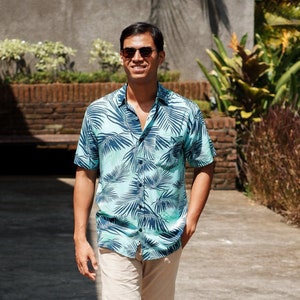 Pode incluir: Uma pessoa vestindo uma camisa de manga curta com estampa de folhas tropicais em tons de azul e verde, combinada com shorts c&aacute;qui e sapatos de barco de couro marrom. A roupa &eacute; adequada para climas quentes.