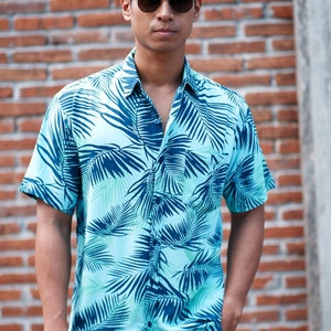 Pode incluir: Uma camisa azul clara com um padr&atilde;o de folhas tropicais em tons de verde-azulado e azul marinho. A camisa de manga curta &eacute; usada com cal&ccedil;as c&aacute;qui e &oacute;culos de sol. O fundo &eacute; uma parede de tijolos.