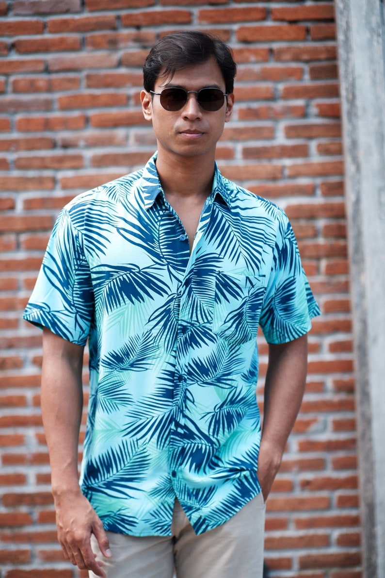 Pode incluir: Uma camisa azul clara com um padr&atilde;o de folhas tropicais em tons de verde-azulado e azul marinho. A camisa de manga curta &eacute; usada com cal&ccedil;as c&aacute;qui e &oacute;culos de sol. O fundo &eacute; uma parede de tijolos.