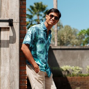 Pode incluir: Uma pessoa vestindo uma camisa turquesa com estampa de folhas de palmeira e shorts c&aacute;qui. A camisa est&aacute; aberta na gola. A pessoa est&aacute; usando &oacute;culos de sol e sorrindo. O fundo inclui uma parede de tijolos e palmeiras.
