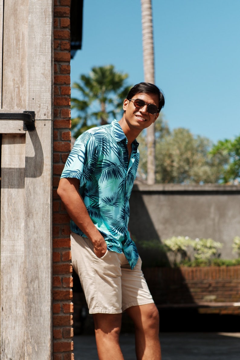 Pode incluir: Uma pessoa vestindo uma camisa turquesa com estampa de folhas de palmeira e shorts c&aacute;qui. A camisa est&aacute; aberta na gola. A pessoa est&aacute; usando &oacute;culos de sol e sorrindo. O fundo inclui uma parede de tijolos e palmeiras.