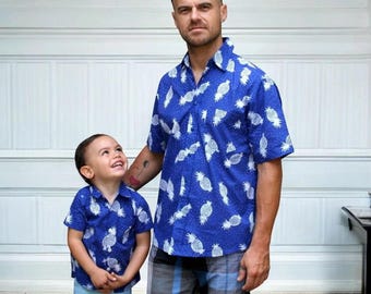 Könnte beinhalten: Ein Vater und sein Sohn tragen passende blaue Hemden mit kurzen Ärmeln und einem weißen Ananasdruck. Die Hemden haben einen Knopfleistenverschluss und einen Kragen. Der Vater trägt eine blaue und graue karierte Shorts. Der Sohn trägt eine blaue Shorts.
