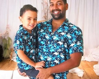 Könnte beinhalten: Ein Vater und sein Sohn tragen passende blau-weiße Hemden mit kurzen Ärmeln und einem Flamingo-Druck. Die Hemden haben einen Knopfkrag und kurze Ärmel.
