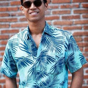 Pode incluir: Camisa azul clara com um padr&atilde;o de folhas tropicais em azul escuro. A camisa de manga curta tem um bolso no peito e &eacute; abotoada. A pessoa na imagem est&aacute; usando &oacute;culos de sol e sorrindo em frente a uma parede de tijolos.