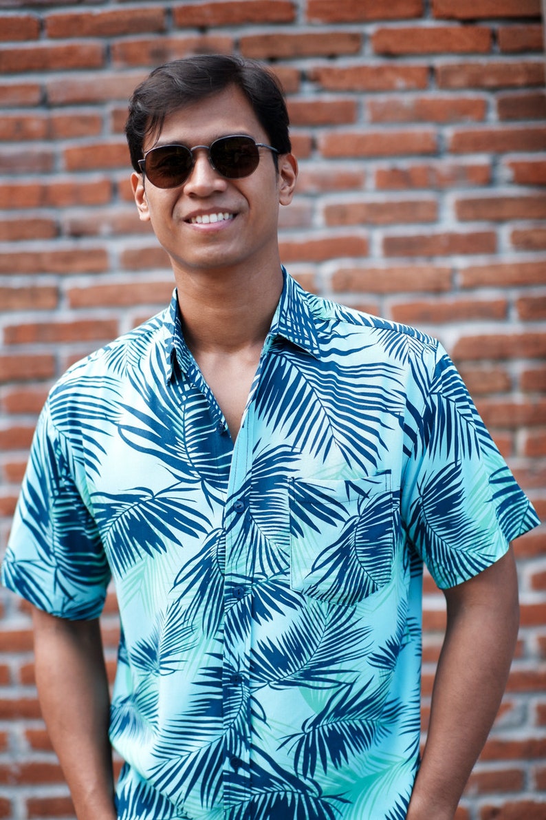 Pode incluir: Camisa azul clara com um padr&atilde;o de folhas tropicais em azul escuro. A camisa de manga curta tem um bolso no peito e &eacute; abotoada. A pessoa na imagem est&aacute; usando &oacute;culos de sol e sorrindo em frente a uma parede de tijolos.