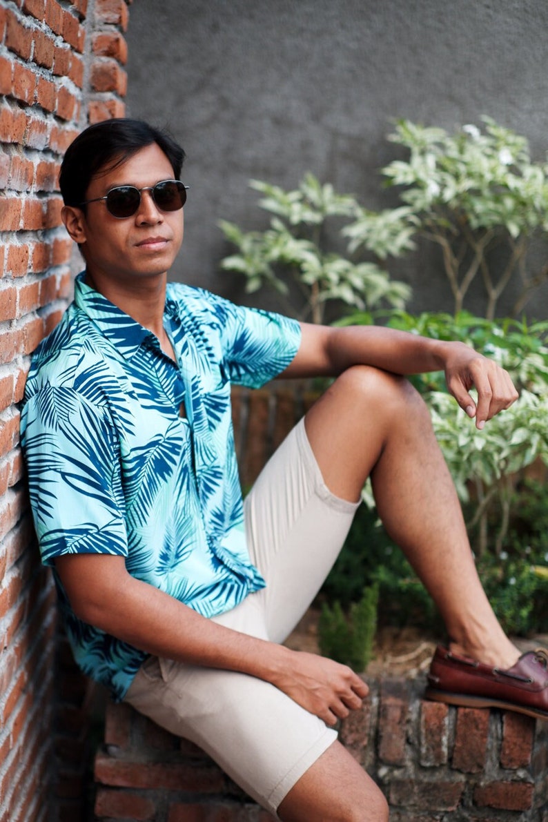 Pode incluir: Uma pessoa vestindo uma camisa azul clara com estampa de folhas tropicais, shorts c&aacute;qui e sapatos de barco de couro marrom. A pessoa est&aacute; usando &oacute;culos de sol.