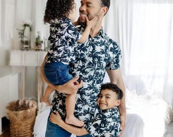 Könnte beinhalten: Ein Vater und seine beiden Söhne tragen passende weiße und schwarze Hemden mit Palmenprint. Der Vater hält seinen jüngsten Sohn im Arm, während der ältere Sohn neben ihm steht. Alle drei lächeln und schauen sich an.