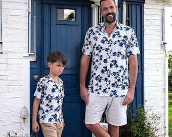 Könnte beinhalten: Ein Vater und sein Sohn tragen passende weiße Hemden mit kurzen Ärmeln und einem blauen Palmen-Druck. Der Vater lächelt und der Sohn schaut mit ernster Miene in die Kamera. Sie stehen vor einer blauen Tür.