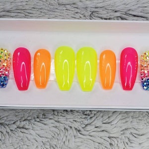 May include: A set of ten neon-colored false nails with a rainbow gradient. The nails are long and oval-shaped. Two of the nails are decorated with clear rhinestones.