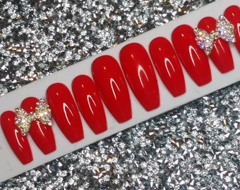May include: Set of ten red coffin-shaped press-on nails with a clear base. Each nail is decorated with a single rhinestone bow.