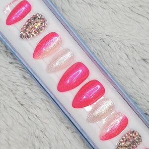 Peut inclure: Un ensemble de dix faux ongles en pression avec un d&eacute;grad&eacute; de rose et de blanc, des paillettes et des strass.