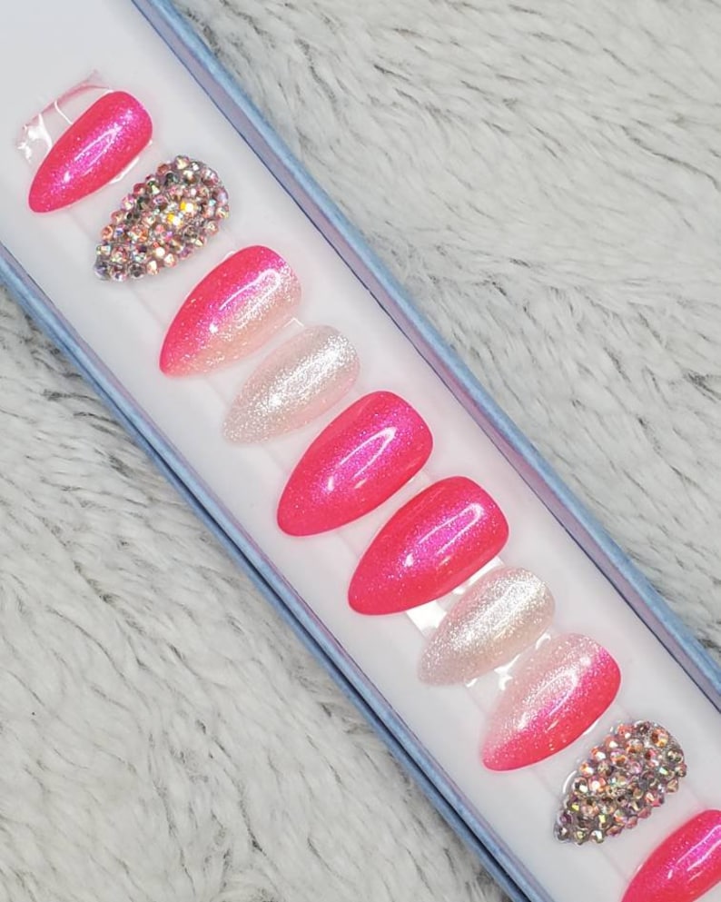 Peut inclure: Un ensemble de dix faux ongles en pression avec un d&eacute;grad&eacute; de rose et de blanc, des paillettes et des strass.