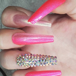 Peut inclure: Longues pointes d'ongles roses &agrave; paillettes avec une rang&eacute;e de strass iris&eacute;s sur un ongle.
