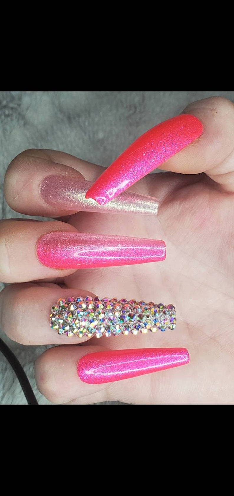Peut inclure: Longues pointes d'ongles roses &agrave; paillettes avec une rang&eacute;e de strass iris&eacute;s sur un ongle.