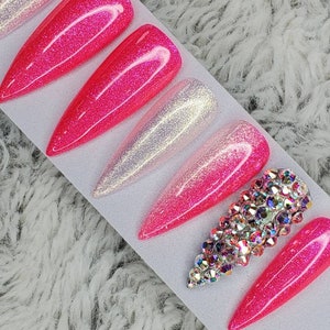 Peut inclure: Un ensemble de dix faux ongles avec un design ombr&eacute; rose et blanc. Les ongles sont longs et pointus, et certains ont des paillettes. Un ongle a un gros groupe de strass.