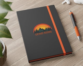May include: Black notebook with an orange elastic closure. The notebook has a graphic design of a mountain range with a sunset behind it. The text "GRAND RISING" is printed below the graphic.