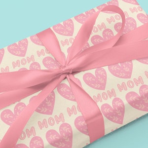 May include: A gift wrapped in cream paper with a pink ribbon. The paper has a repeating pattern of pink hearts with the word "MOM" inside each heart.