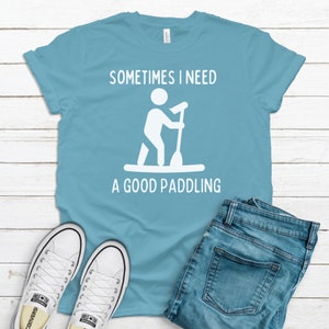 May include: A light blue t-shirt with the text "Sometimes I need a good paddling" and a white silhouette of a person paddleboarding. The shirt is laid flat on a white wooden surface with a pair of white sneakers and blue jeans.