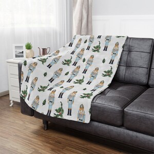 May include: White plush throw blanket with a repeating pattern of nutcracker soldiers and holly sprigs. The blanket is folded and draped over a black leather couch.