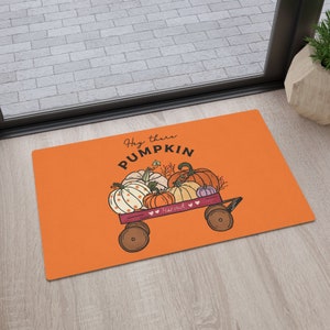 Może przedstawiać: Pomarańczowa wycieraczka z napisem "Hey there PUMPKIN" i czerwonym wozem wypełnionym dyniami. Wóz ma napis "Harvest" z boku.