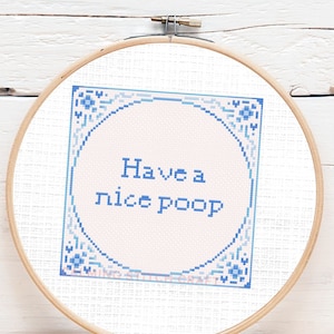 Op de afbeelding: Een witte borduurring met een blauw en wit kruissteekontwerp. Het ontwerp heeft een roze cirkel met de tekst "Have a nice poop" in blauw.