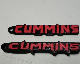May include: Two black and red "Cummins" logo emblems. The word "Cummins" is in red, with a black outline. The emblems are on a light gray surface. A black pen is in the background.