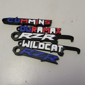 May include: A collection of black 3D-printed keychains, each bearing a different name: "Cummins", "Duramax", "RZR", and "Wildcat". The text is in various colours, including white, red, and blue.