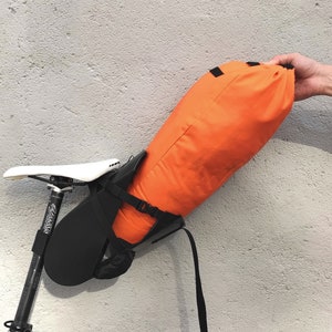 Op de afbeelding: Een feloranje waterdichte fietssaddle tas bevestigd aan een zwart zadel met een zwarte band. De tas wordt vastgehouden door een hand.