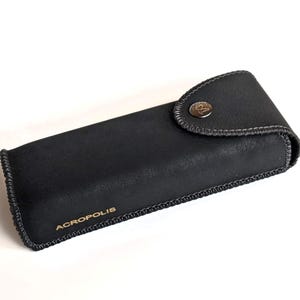 May include: A black rectangular case with the word "ACROPOLIS" in gold lettering. The case has a button closure and a stitched border. The case is likely for eyeglasses or sunglasses.
