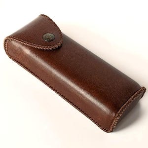 May include: A brown leather case with a button closure. The case has a stitched edge and is made of a soft, supple leather.