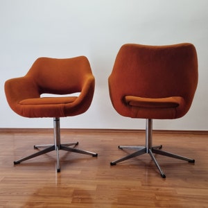 May include: Two orange swivel chairs with a mid-century modern design. The chairs have a rounded back and seat, with a brushed metal base featuring a five-star design. The chairs are set against a white wall and a wood floor.