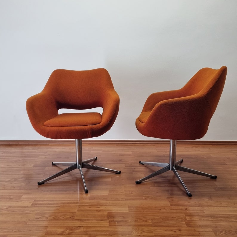 May include: Two orange swivel chairs with a mid-century modern design. The chairs have a rounded seat and back with armrests. They sit on a silver, star-shaped base. The chairs are placed on a wood floor against a white wall.